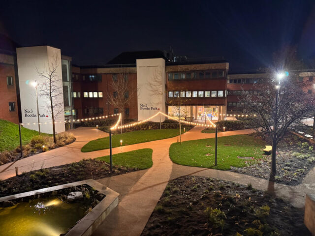 Booths Park courtyard after dark, with illuminated paths, lawns and planted beds.